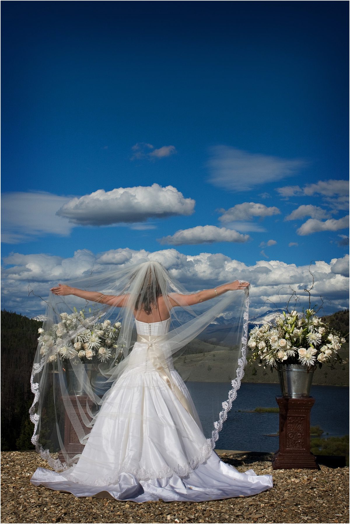 bridal portrait, bride at ceremony altar overlooking mountain lake, puffy white clouds and blue sky, holding out the veil,C Lazy U Ranch, Granby, Colorado Wedding Photography, Mountain Wedding Photographer, bride alone on wedding day